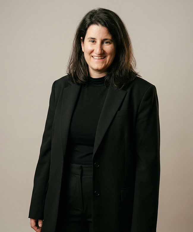 Phygital Group A person with shoulder-length dark hair wearing a black blazer and black shirt stands facing the camera and smiling against a plain light background, embodying the professionalism of a leading Marketing Agency. Full Service Marketing Agency