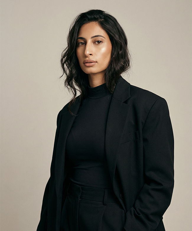 Phygital Group A person with wavy dark hair wearing a black blazer and black top stands against a plain beige background, embodying the confident, modern spirit of a leading marketing agency while looking directly at the camera. Full Service Marketing Agency