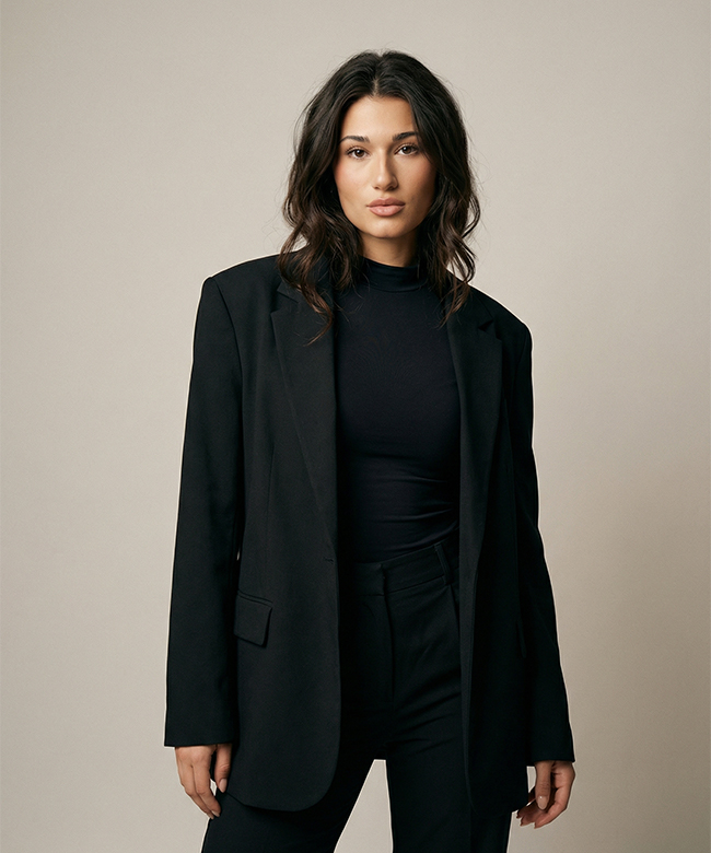 Phygital Group A woman with wavy dark hair, representing a growth agency, wears a black blazer over a black top and pants, standing against a plain beige background. Full Service Marketing Agency