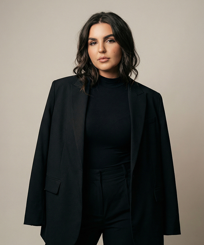 Phygital Group A woman with long, dark hair, dressed in a black blazer with matching top and pants, stands confidently against a plain light-colored background—perfect for showcasing your Growth Agency’s professional team. Full Service Marketing Agency