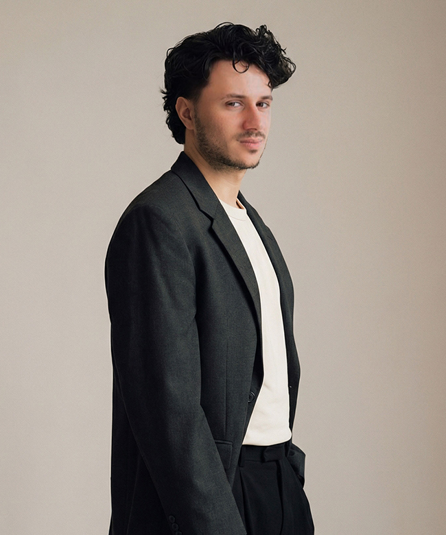 Phygital Group A man with wavy dark hair wearing a black blazer, white t-shirt, and black pants stands against a plain light background, exuding the confident presence of a Growth Agency professional as he looks at the camera. Full Service Marketing Agency