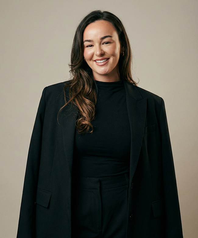 Phygital Group A woman with long brown hair, in a black blazer and top, stands against a plain light background, smiling—reflecting the confident professionalism of a leading Marketing Agency. Full Service Marketing Agency