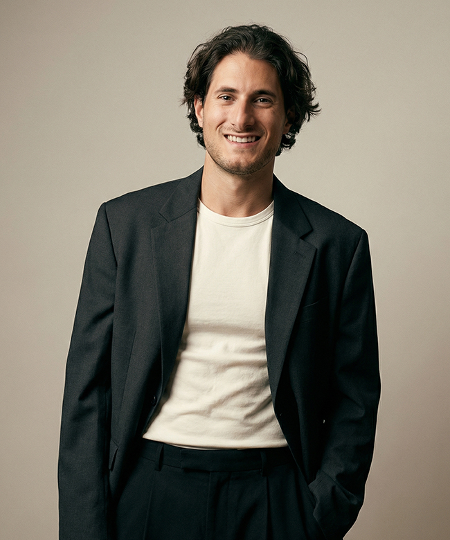 Phygital Group A man with medium-length dark hair wears a black suit jacket over a white t-shirt, standing and smiling against a plain beige background—reflecting the approachable style of a modern Marketing Agency. Full Service Marketing Agency