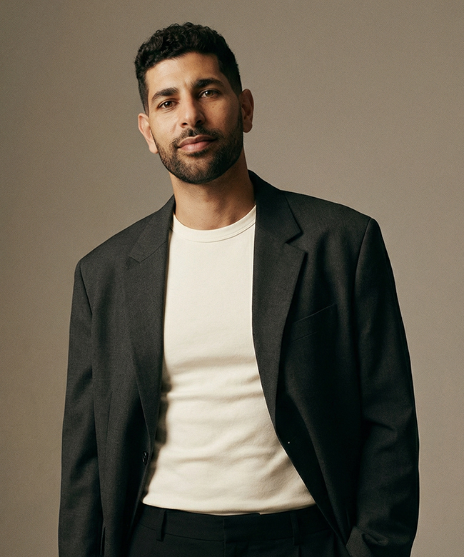 Phygital Group Man with short curly hair and a beard wearing a dark blazer over a light-colored t-shirt, standing against a plain background, exuding the confident professionalism of someone from a leading Growth Agency. Full Service Marketing Agency