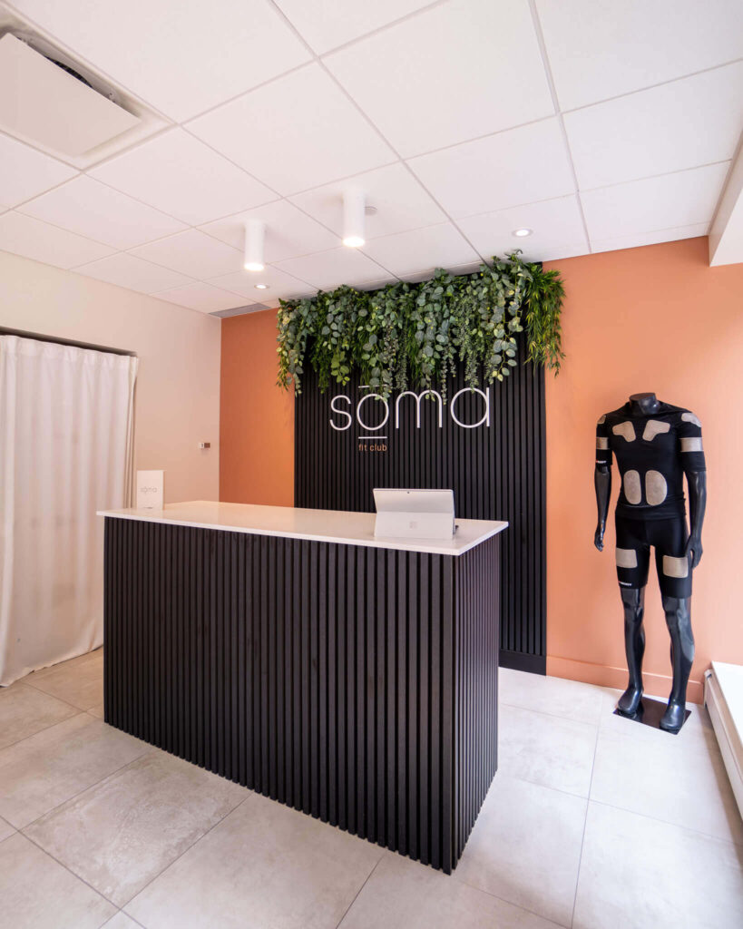 Phygital Group Reception desk with a laptop in front of a sign reading "soma," green hanging plants above, and a mannequin wearing a black suit with white patches to the right—perfect for an innovative marketing agency space. Full Service Marketing Agency