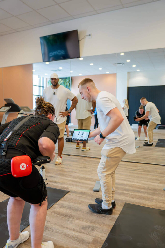 Phygital Group A trainer from a marketing agency shows a fitness screen to a person wearing EMS gear in a gym, with two others observing and a mirrored wall reflecting the scene. Full Service Marketing Agency